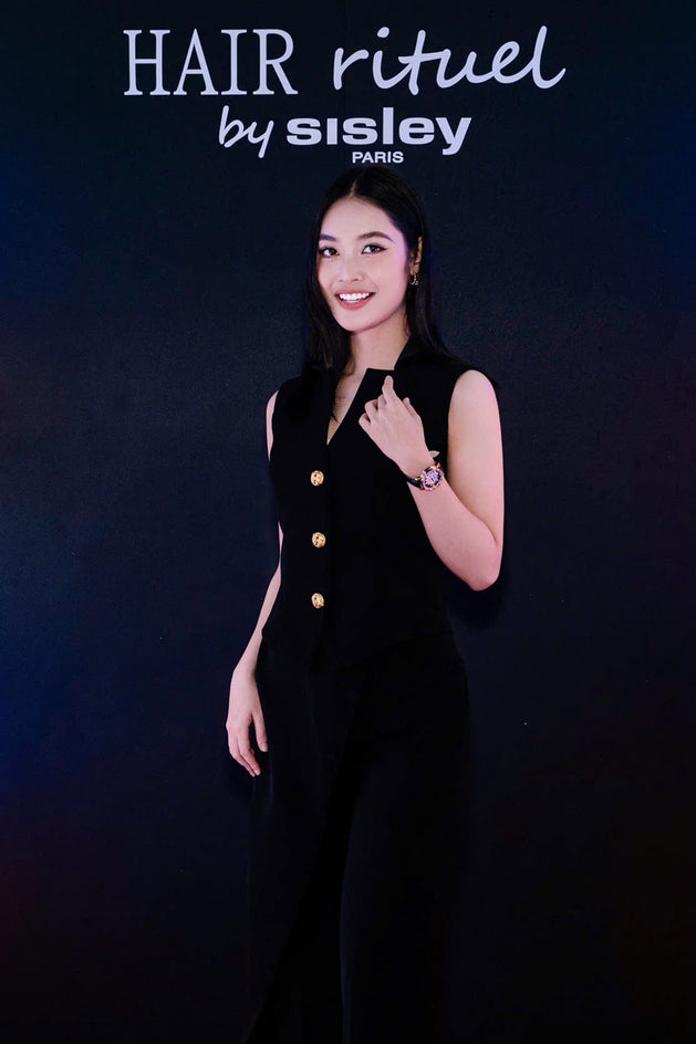A woman stands against a dark blue background wearing a black sleeveless jumpsuit with gold buttons.